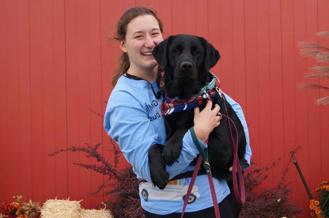 Grace, 微笑着，穿着一件蓝色的运动衫, 在一个红色的谷仓前，抱着一只戴着红蓝色大手帕的黑色拉布拉多犬.
